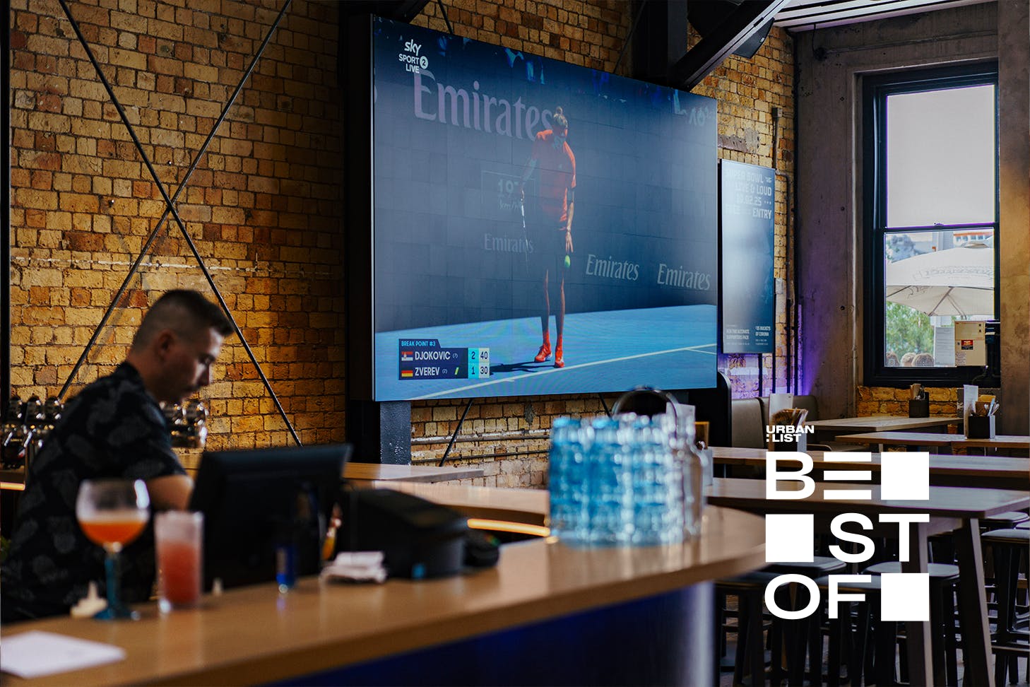 A bartender is busy behind the bar while a sports game plays on a massive screen at Tyler St Sport.